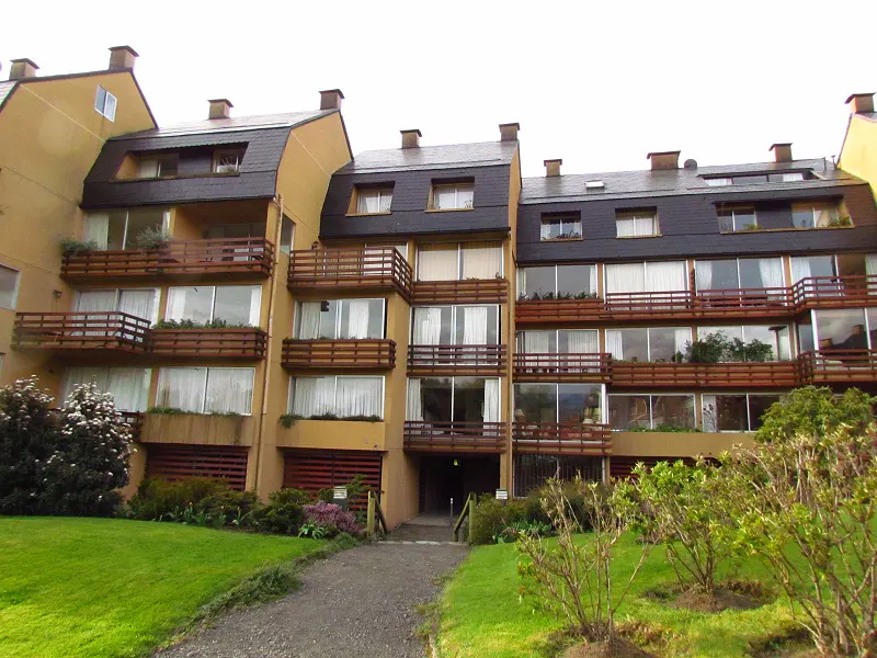 A tan apartment building with dark roofs and wooden balconies, surrounded by green grass and plants.