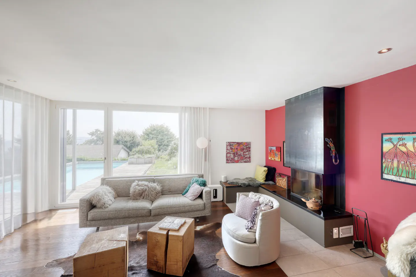 Bright living room with a gray sofa, white chair, and a modern fireplace against a red wall. Large windows overlook a pool.