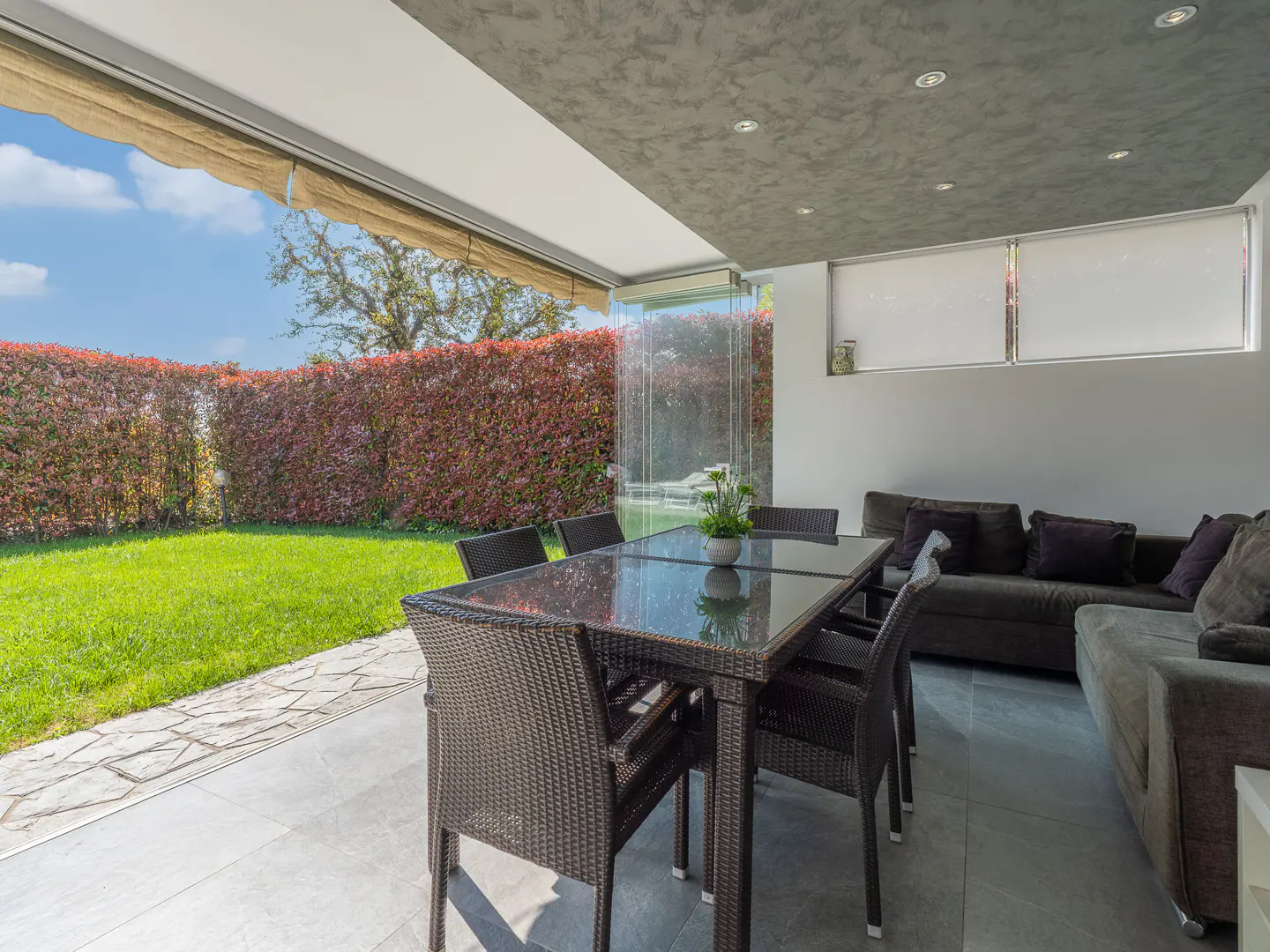 Outdoor patio with a dark wicker table and chairs, a gray sofa, and a view of a green lawn and red hedge.