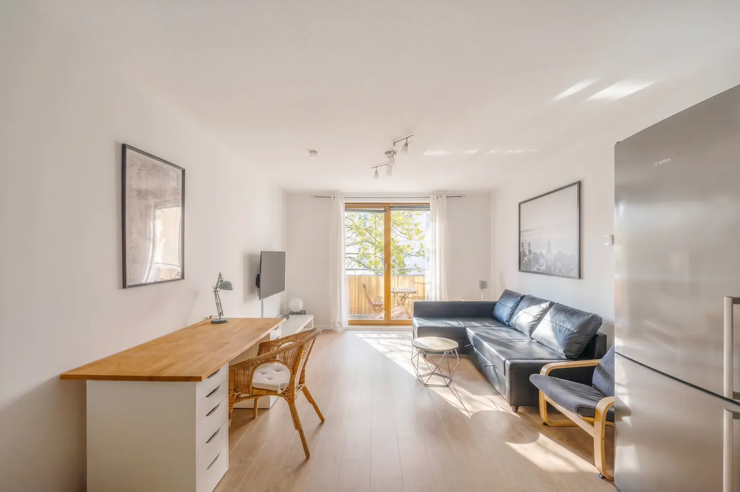 Bright living room with a black leather couch, wood floors, desk, and balcony with a table and chairs.