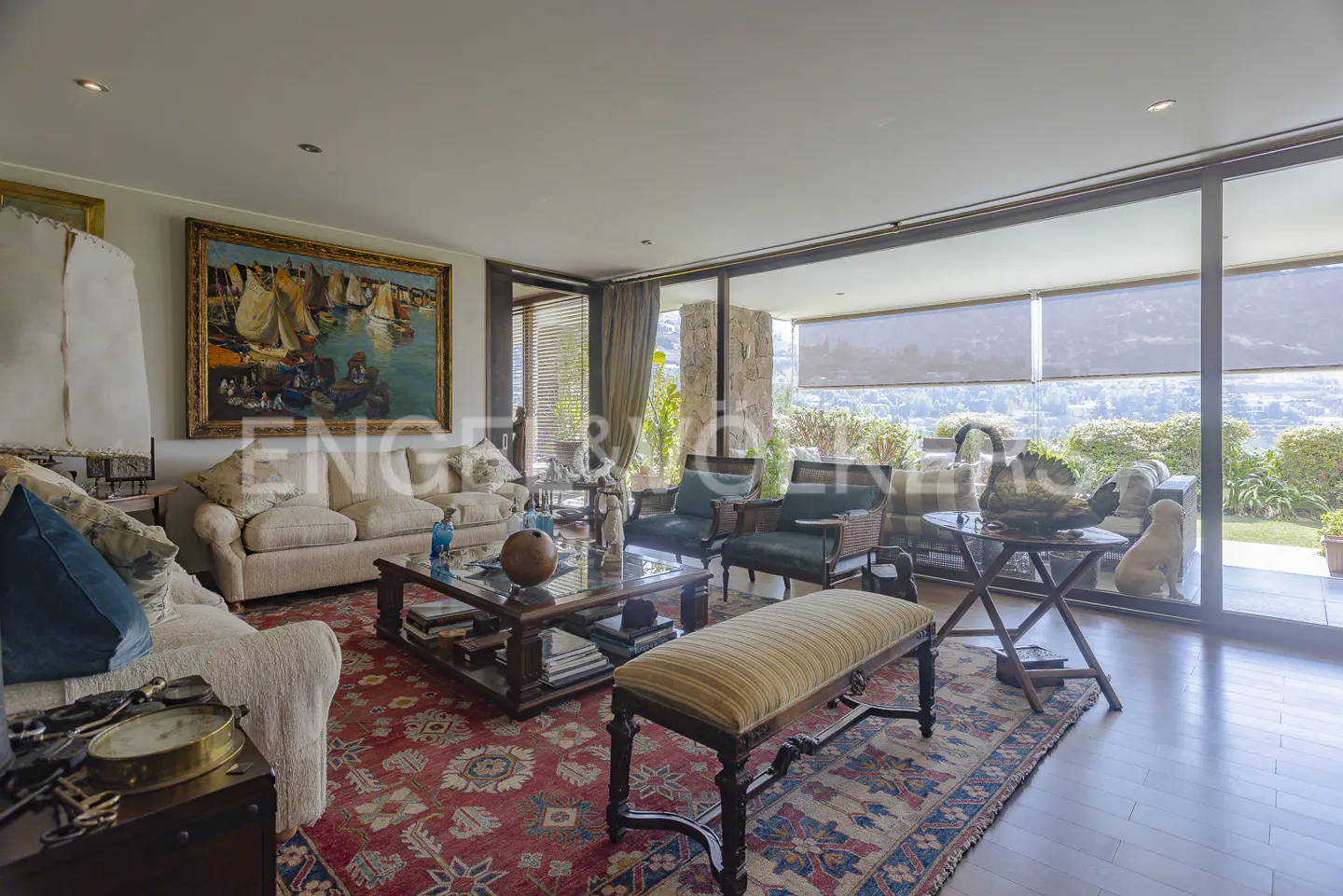 Bright living room with sofas, chairs, and a red patterned rug. Large windows offer a view of greenery. Artwork adorns the walls.