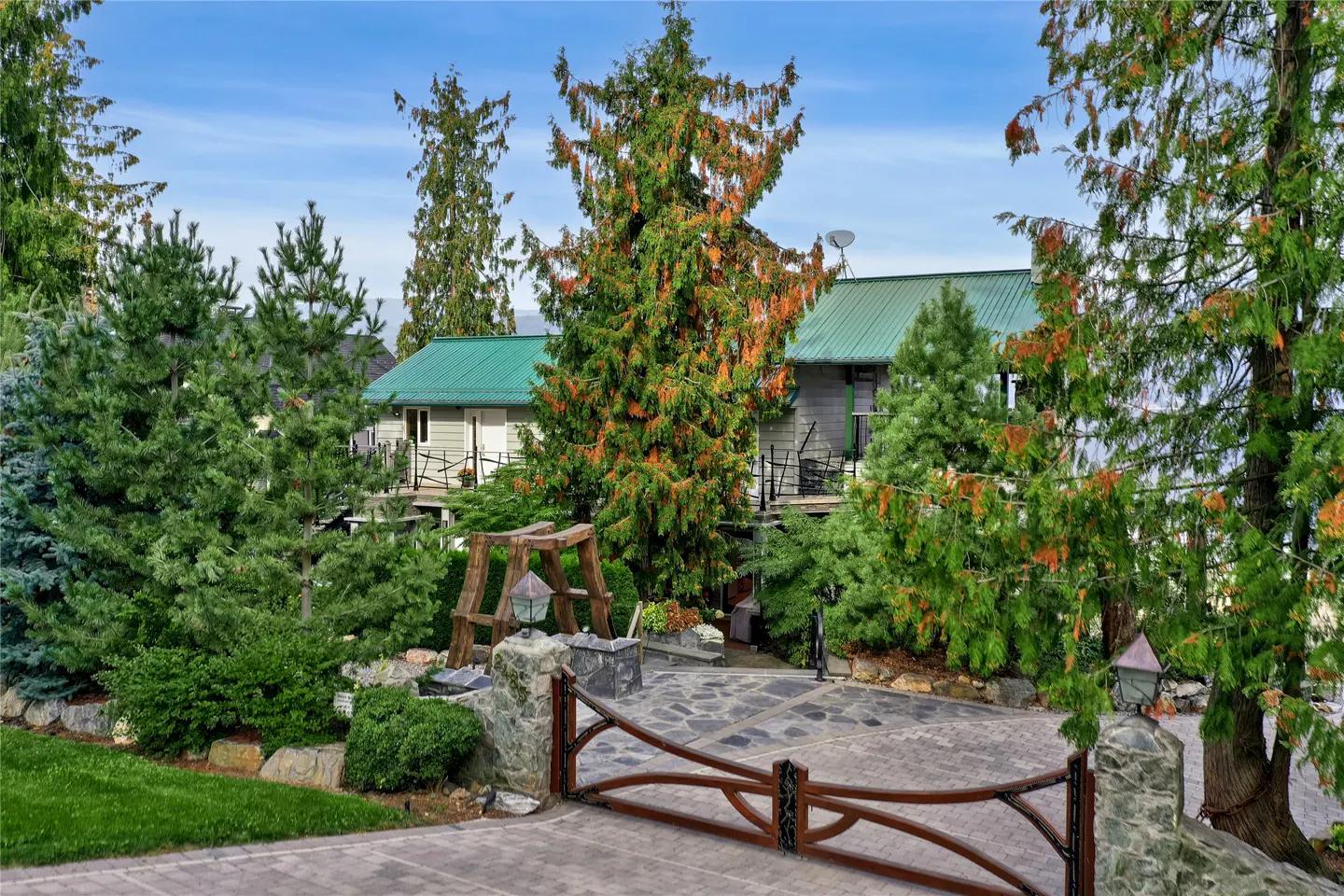 Exterior view of a gray house with a green roof, surrounded by trees and a wooden gate.