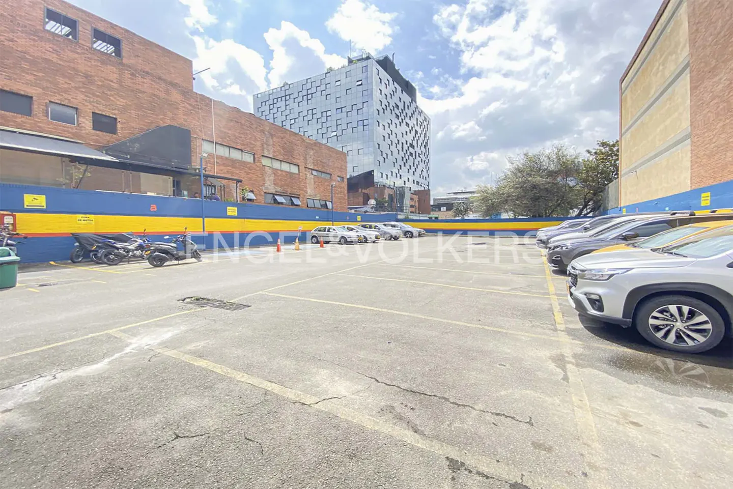 Outdoor parking lot with cars and motorcycles parked against a blue and yellow wall, with a modern building in the background.