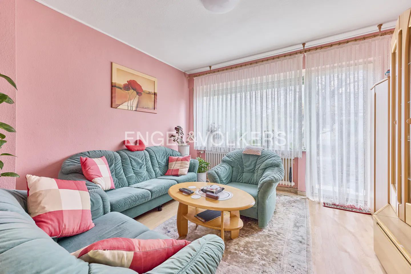 Living room with pink walls, teal sofas with pink pillows, and a light wood coffee table on a patterned rug. Curtains cover the window.
