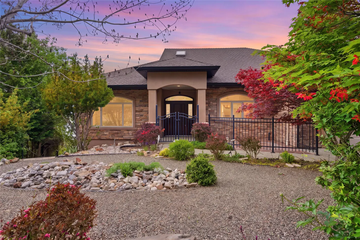 A stone house with a dark roof, arched windows, and a black metal gate is surrounded by green trees and bushes.