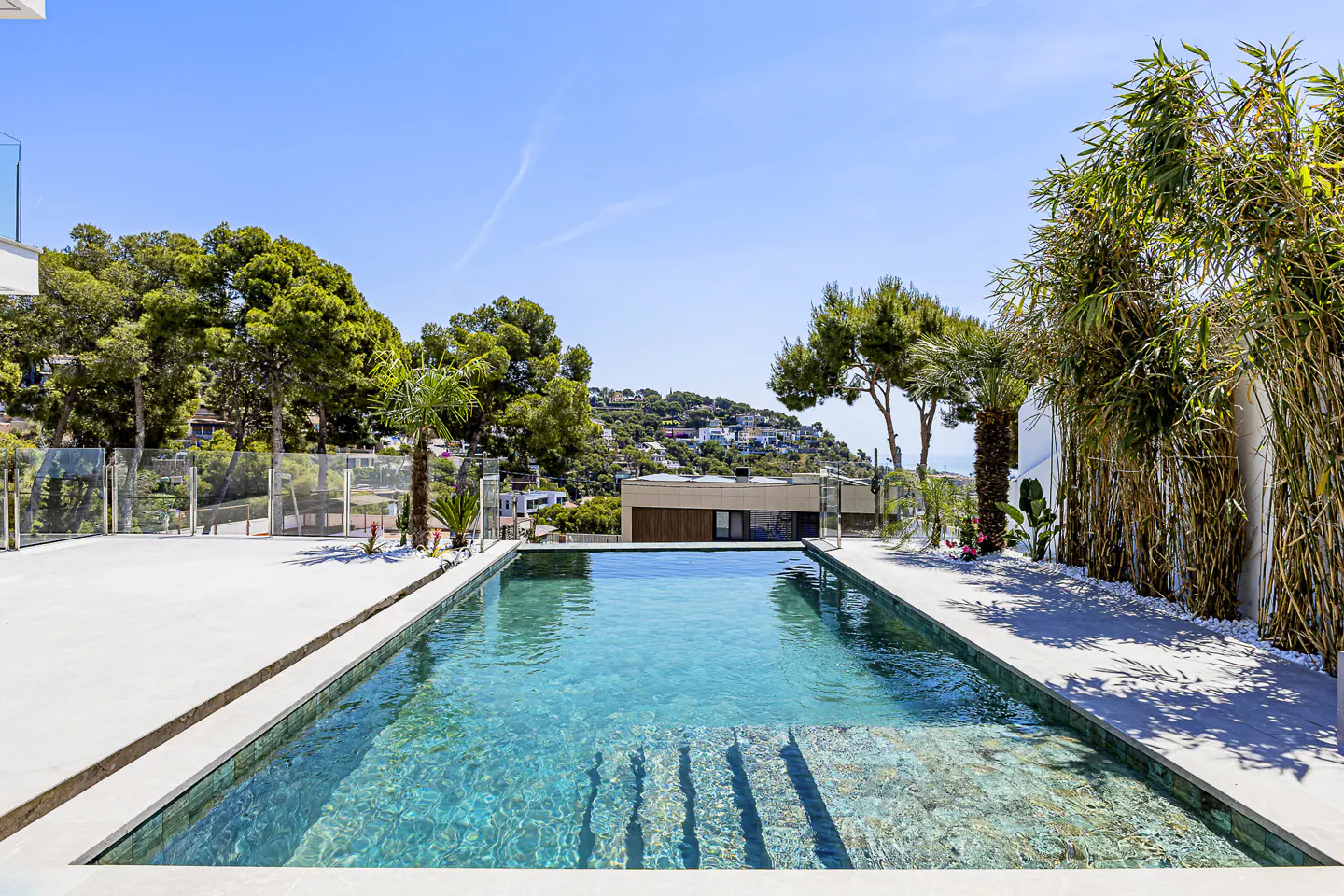 A rectangular turquoise pool with steps, surrounded by a white patio, trees, and a hillside of houses under a blue sky.