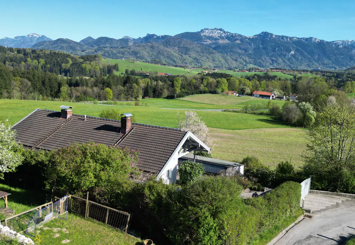 A house with a brown roof sits in front of green fields and mountains under a blue sky.