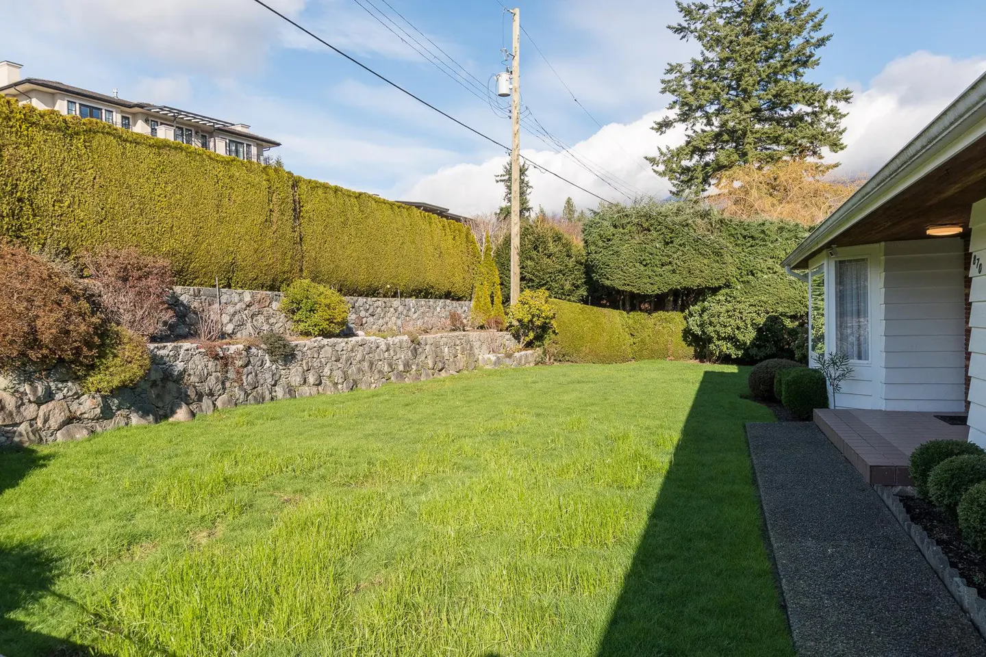 A wide green lawn slopes up to a stone wall and tall hedge. A white house is on the right. Blue sky.