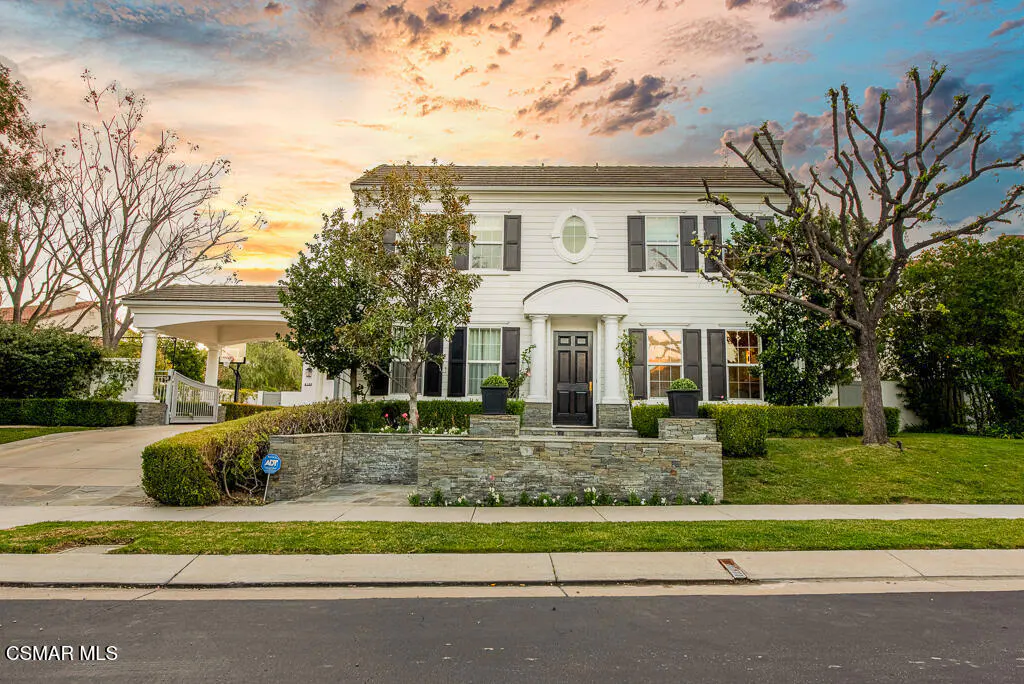 Two-story white house with black shutters and door, stone wall, green lawn, and trees under a colorful sunset sky.