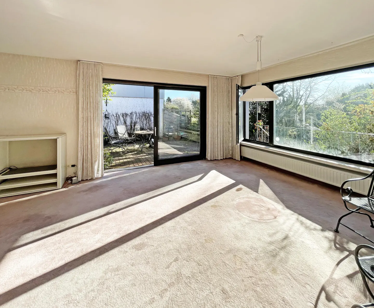 Bright living room with pink carpet, white walls, and large windows. Sliding glass doors lead to a patio with outdoor furniture.