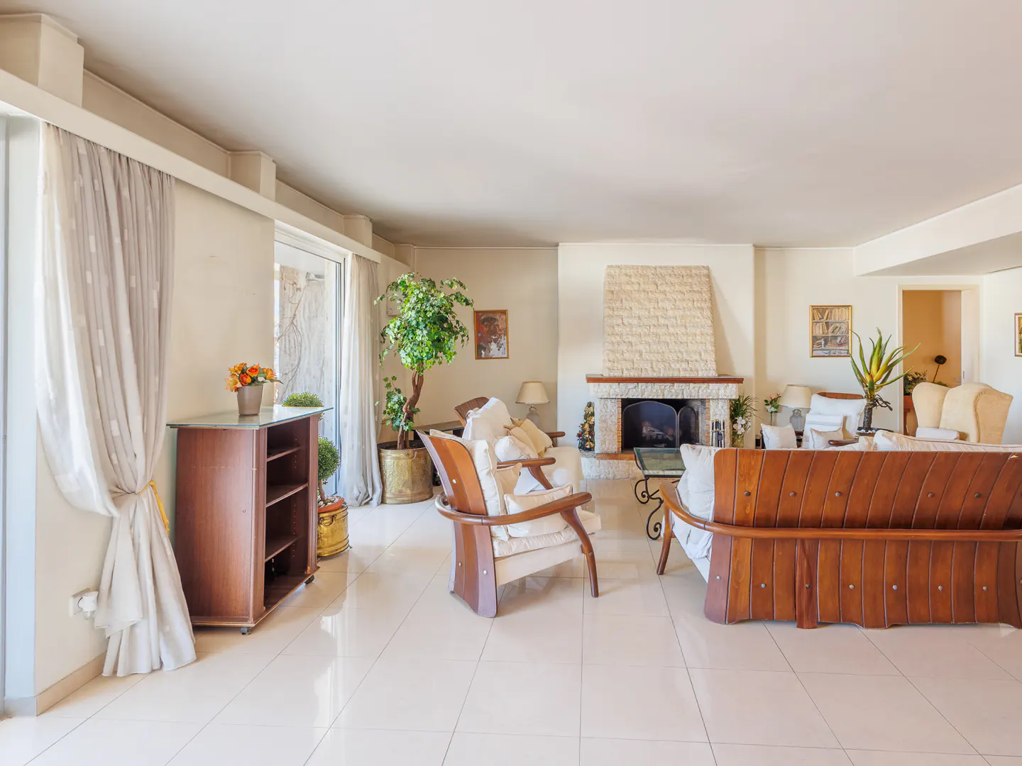 Bright living room with white tile floor, stone fireplace, wood furniture, and large windows with sheer curtains.