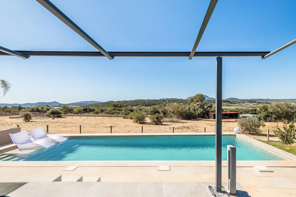 A modern pool with two white lounge chairs sits in turquoise water, under a metal pergola, with a rural landscape in the background.