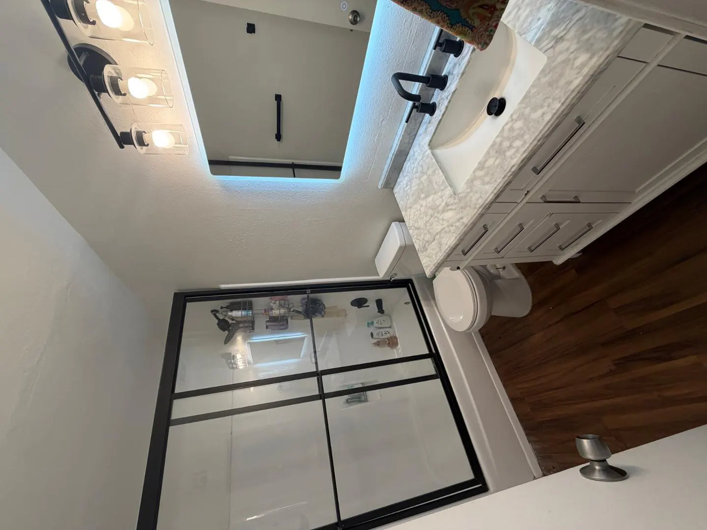 Bathroom with a black-framed glass shower, white vanity with marble top, and wood floors.