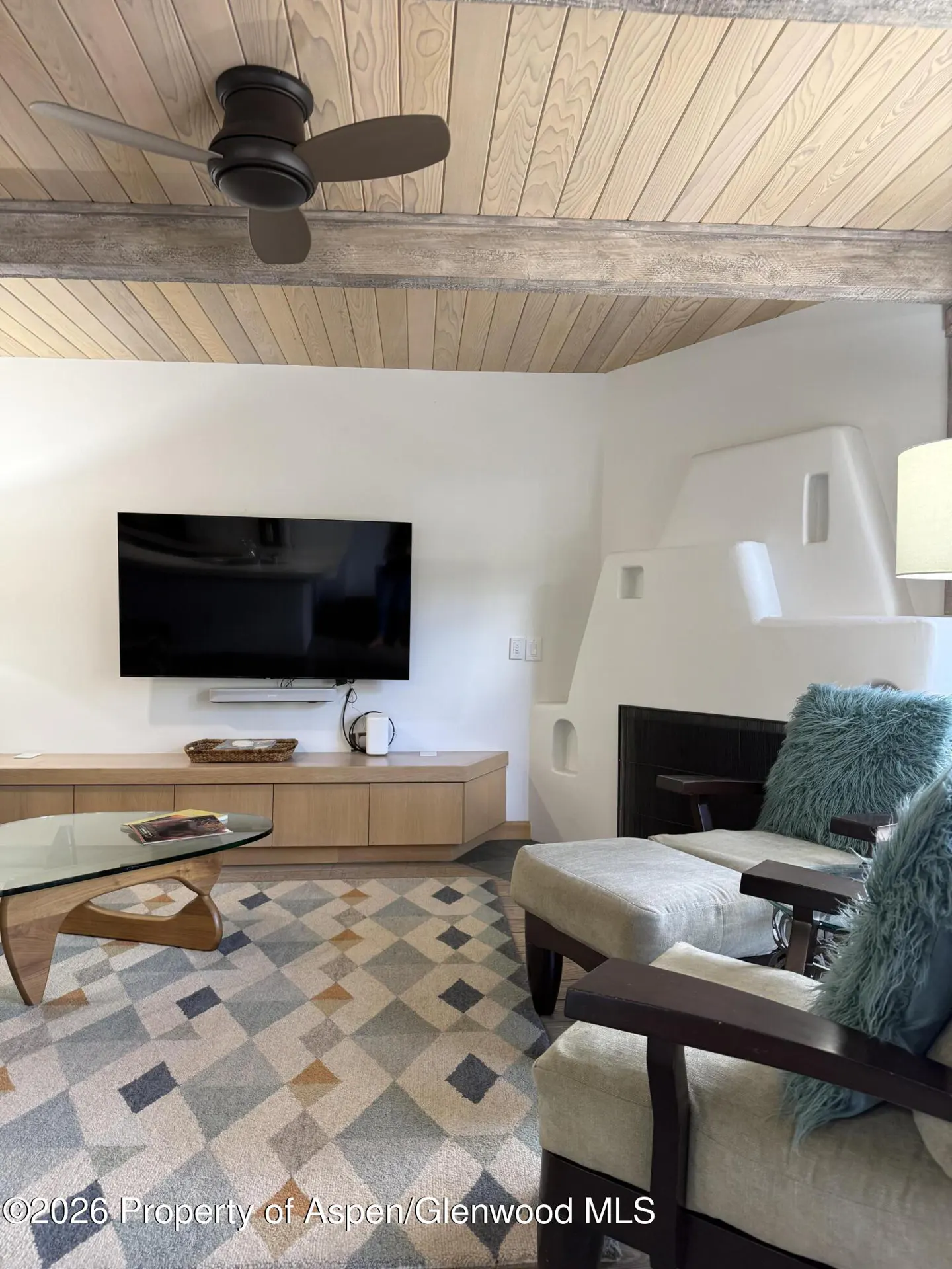 Living room with wood-paneled ceiling, ceiling fan, wall-mounted TV, geometric rug, and two chairs with teal pillows.