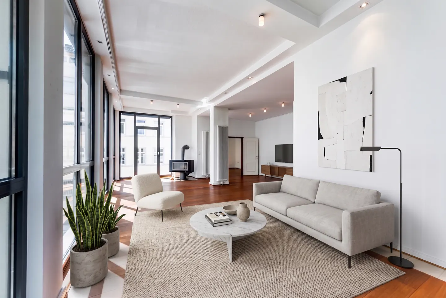 Bright living room with hardwood floors, large windows, and a neutral color palette. A gray sofa, white chair, and marble table furnish the space.