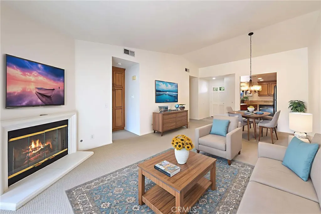 A bright living room with a fireplace, TV, sofa, chairs, and a blue patterned rug. The kitchen and dining area are visible in the background.
