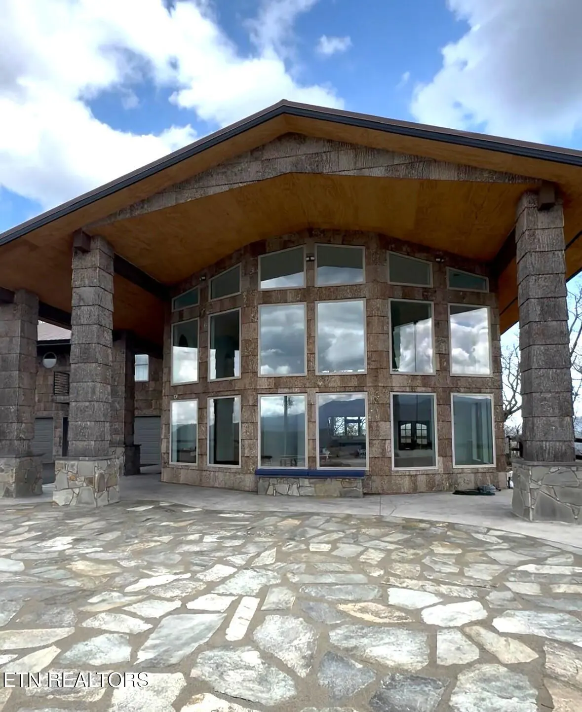 Exterior view of a stone house with a wall of windows and a stone patio under a cloudy sky.