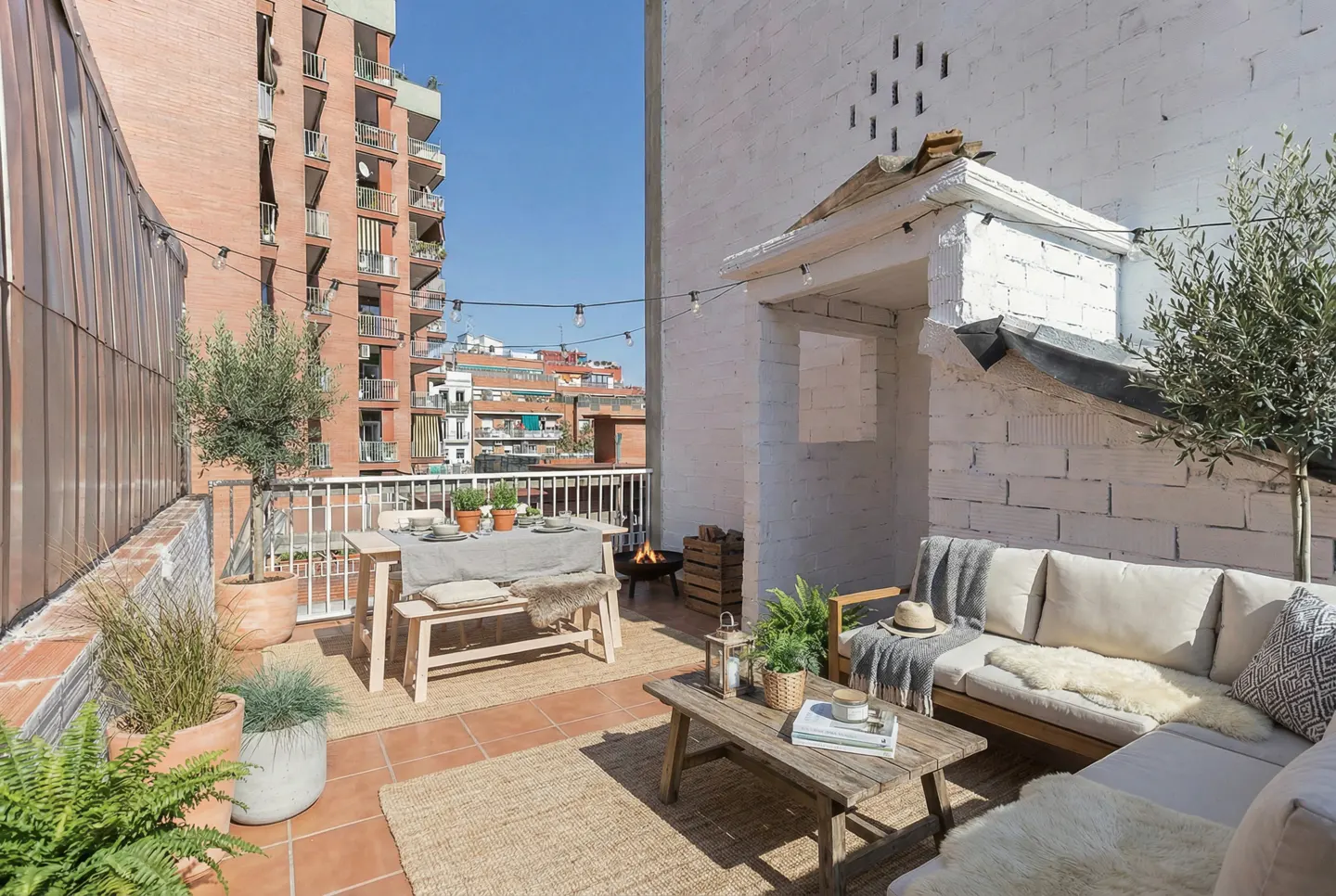 A rooftop patio with a dining table, sofa, coffee table, and potted plants. String lights hang overhead. A brick building is in the background.