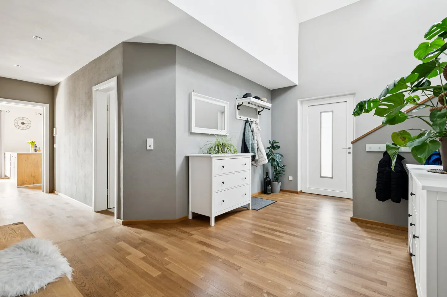 A modern home's entryway with hardwood floors, gray walls, and white trim. A white dresser sits beneath a mirror and coat rack. A large monstera plant adds a touch of green.