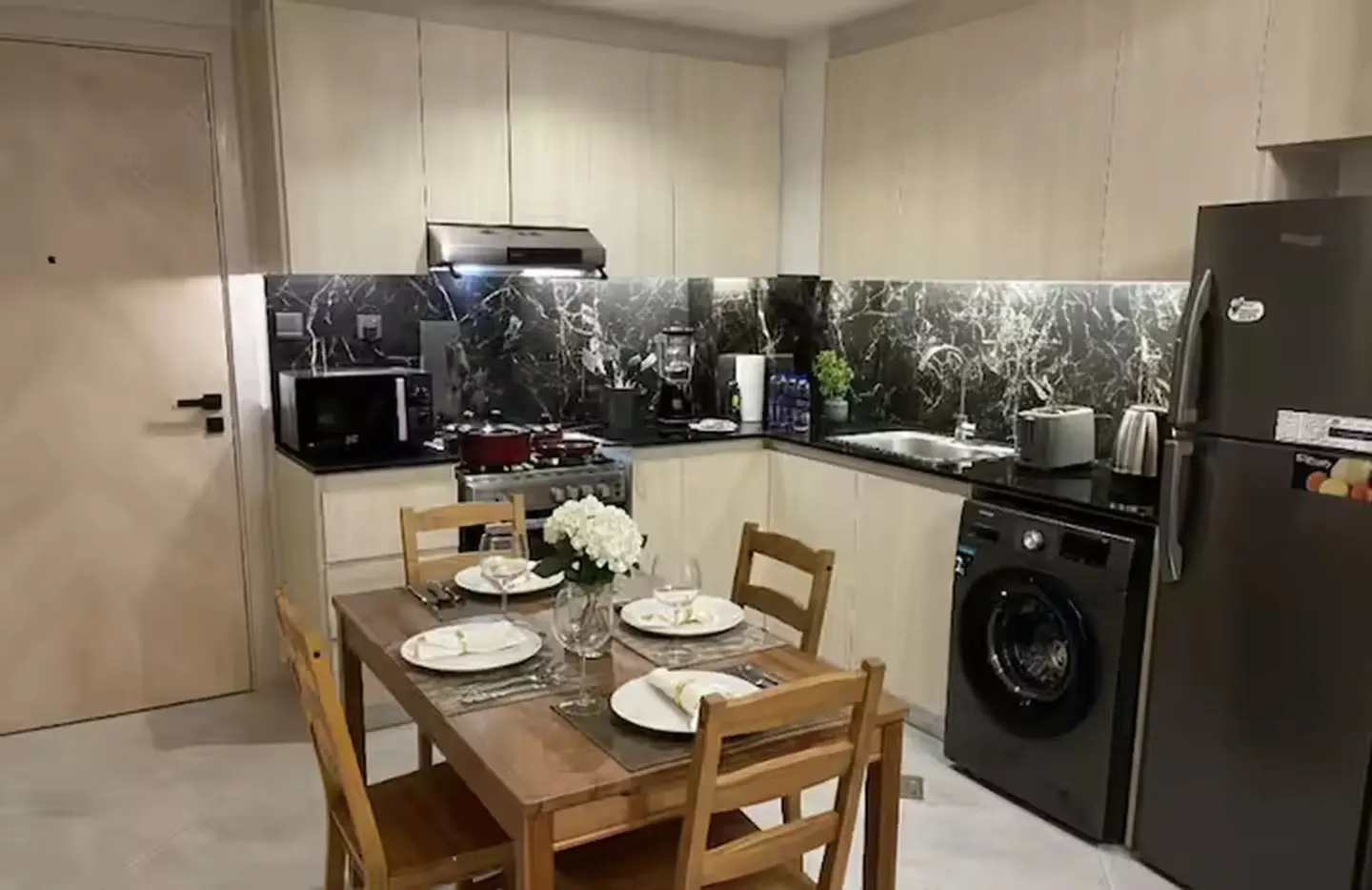 A modern kitchen with light wood cabinets, black marble backsplash, and stainless steel appliances. A wooden dining table with four chairs is set for a meal.