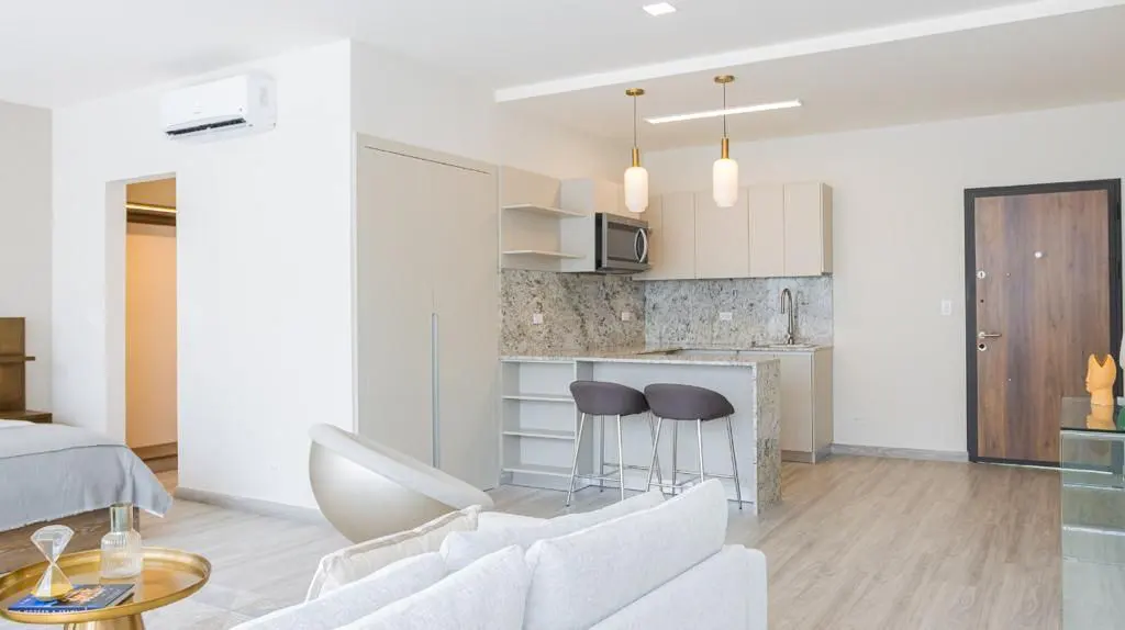 Bright, modern apartment interior with a kitchen island, bar stools, and a white sofa. A bedroom is visible in the background.