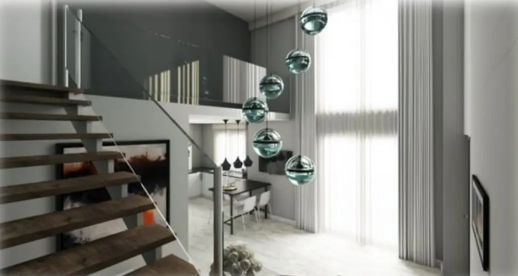 Interior view of a modern home with wooden stairs, glass railing, and hanging globe lights. A kitchen and large window are visible in the background.