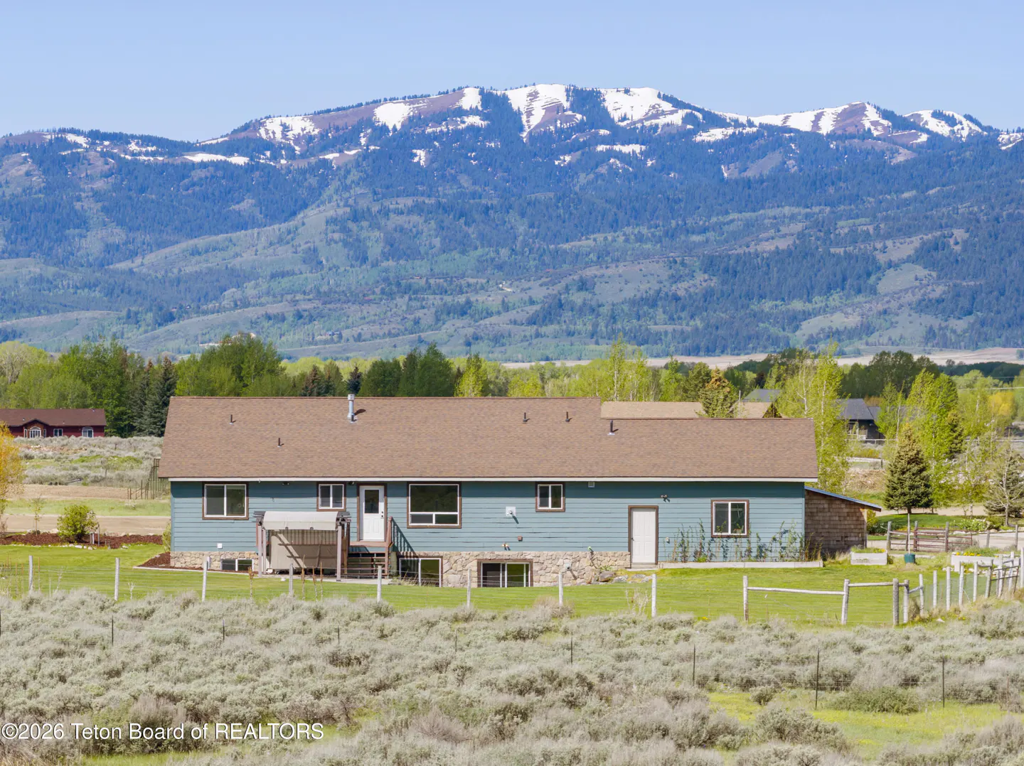 Exterior view of a long, light blue house with a brown roof, set against a backdrop of mountains and trees.