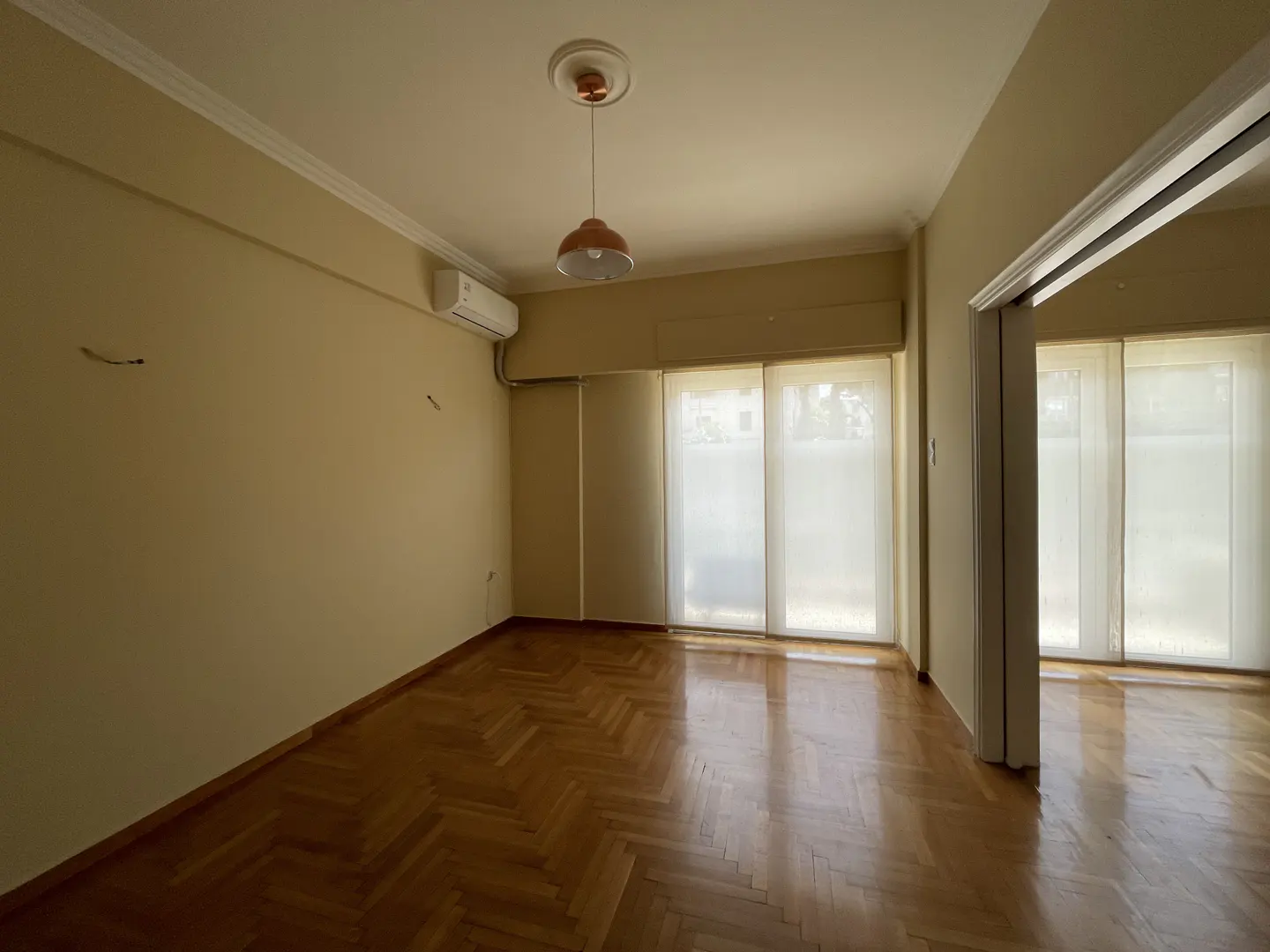 Empty room with parquet floor, beige walls, and white trim. A salmon-colored pendant light hangs from the ceiling. Windows with sheer curtains let in natural light.