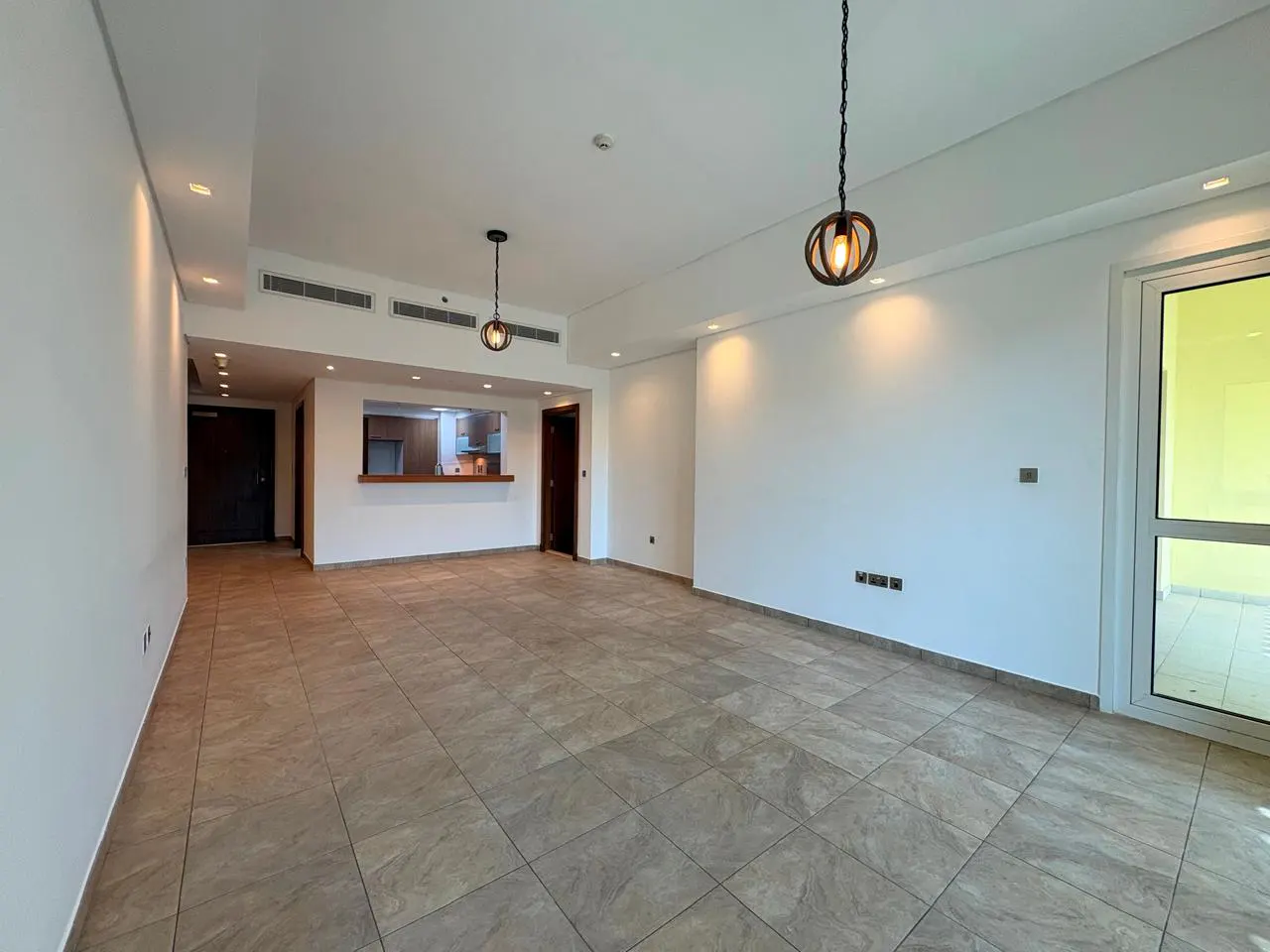 An empty living room with beige tile flooring, white walls, and two hanging pendant lights. A kitchen is visible through a serving hatch.