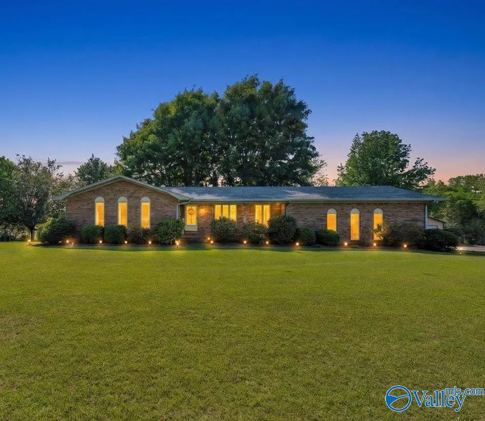 A single-story brick house with a large lawn at dusk. Lights shine from the windows and landscape lighting.