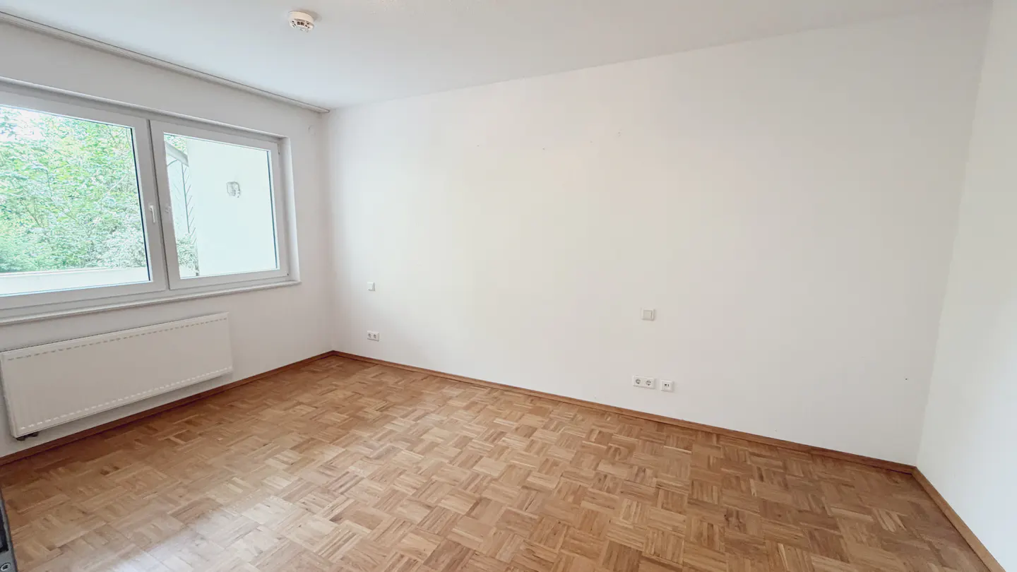 Empty room with parquet floor, white walls, and a window showing green trees outside.