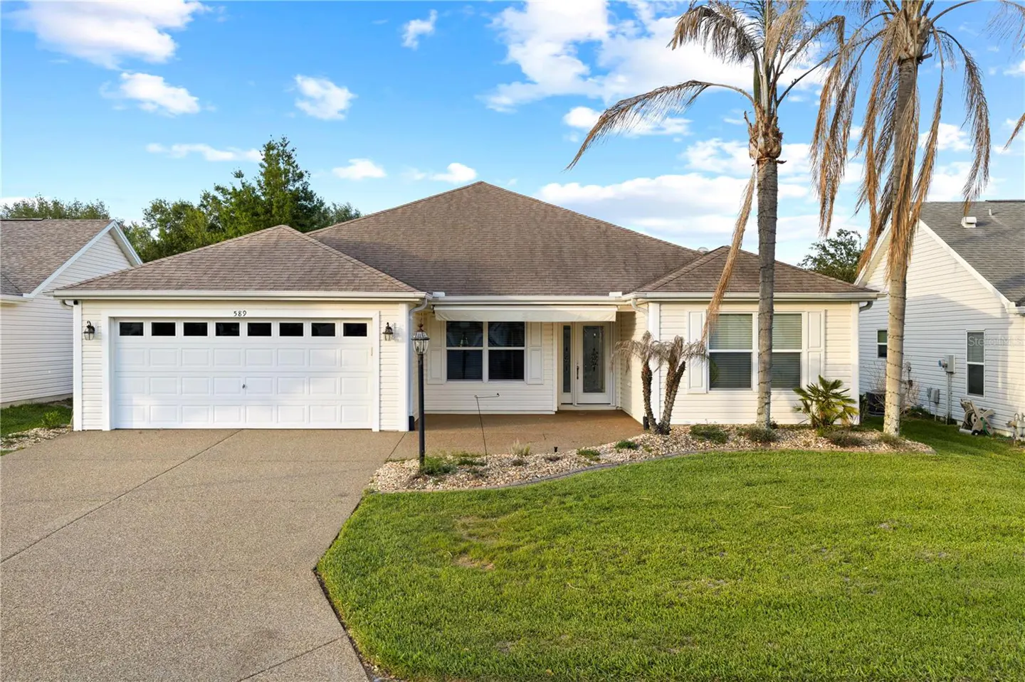 A single-story white house with a brown roof, a two-car garage, and a green lawn. Palm trees stand in the front yard.