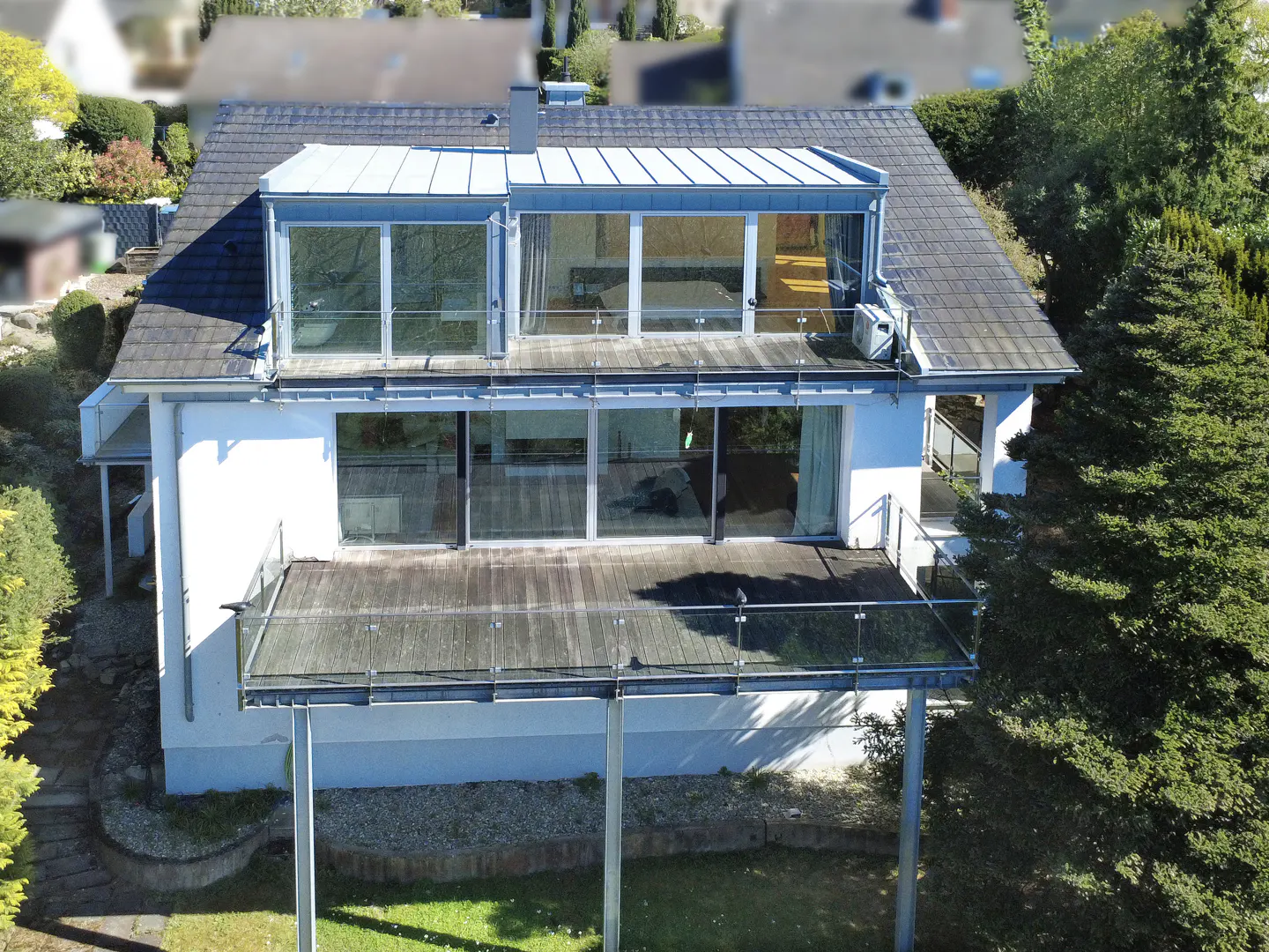 Two-story white house with a gray roof, large windows, and a wooden deck with glass railings. Green trees surround the house.