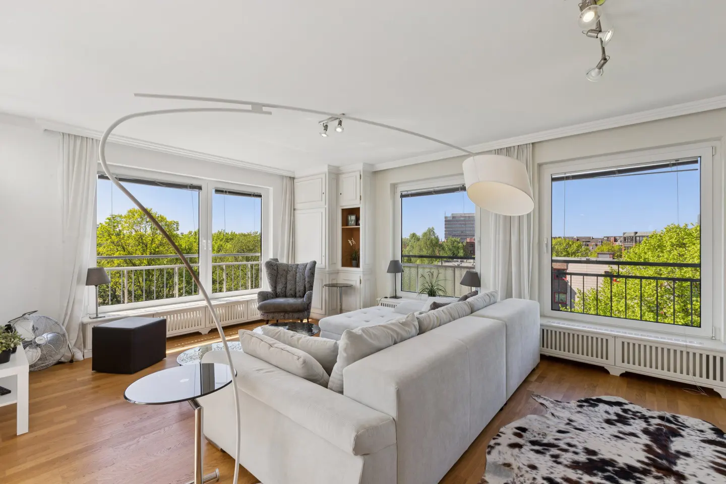 Bright living room with hardwood floors, white sofa, and large windows overlooking green trees. A cowhide rug adds texture.