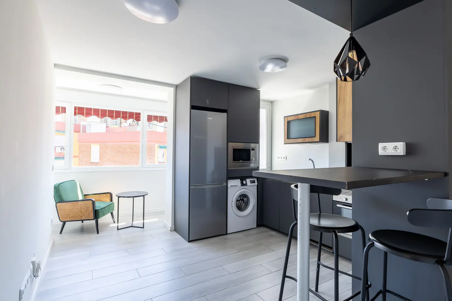 A modern kitchen with gray cabinets, stainless steel appliances, and a black countertop bar with stools. A green chair sits near a window.