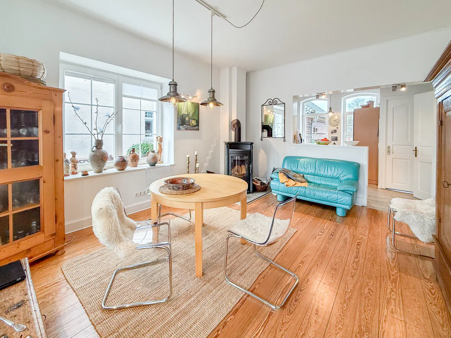 Bright living room with wood floors, a round table with chairs, and a turquoise sofa. A black fireplace and a wooden cabinet add character.