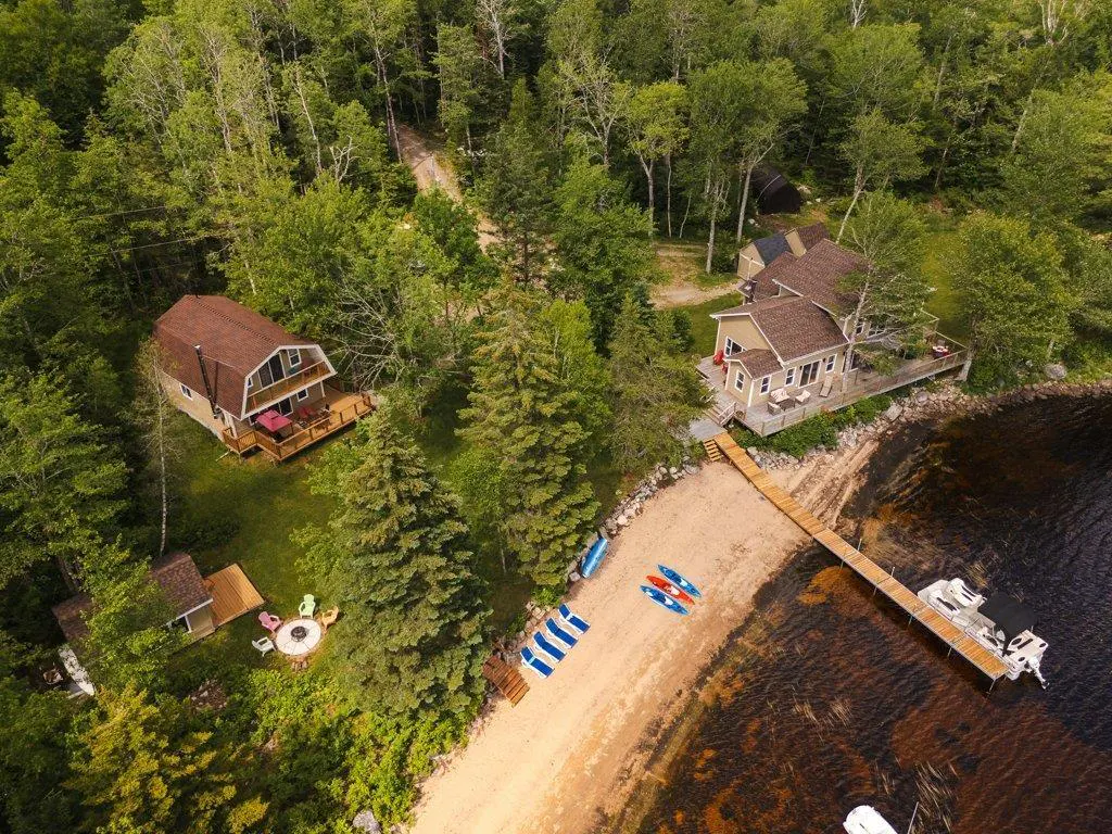 Aerial view of two houses on a lake, surrounded by trees. A sandy beach has kayaks, chairs, and a dock with boats.