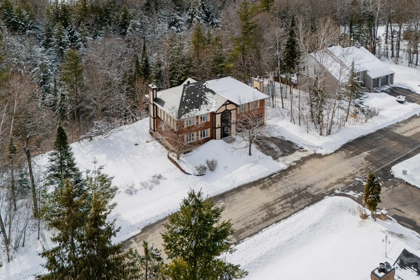 Aerial view of a brick house with a snow-covered roof, surrounded by snow-covered trees and a road.