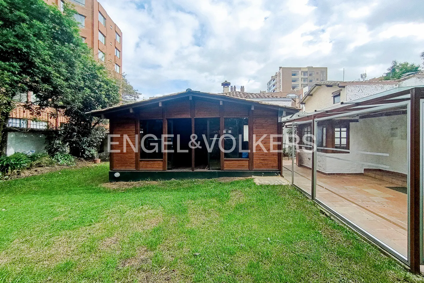 Exterior view of a brown wooden cabin with glass windows, a green lawn, and a glass-enclosed patio.