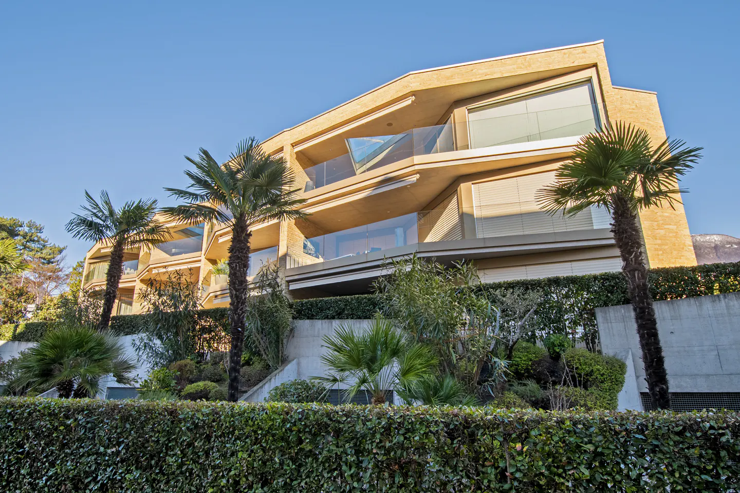 A modern, multi-story tan building with glass balconies and palm trees in front of a green hedge under a blue sky.