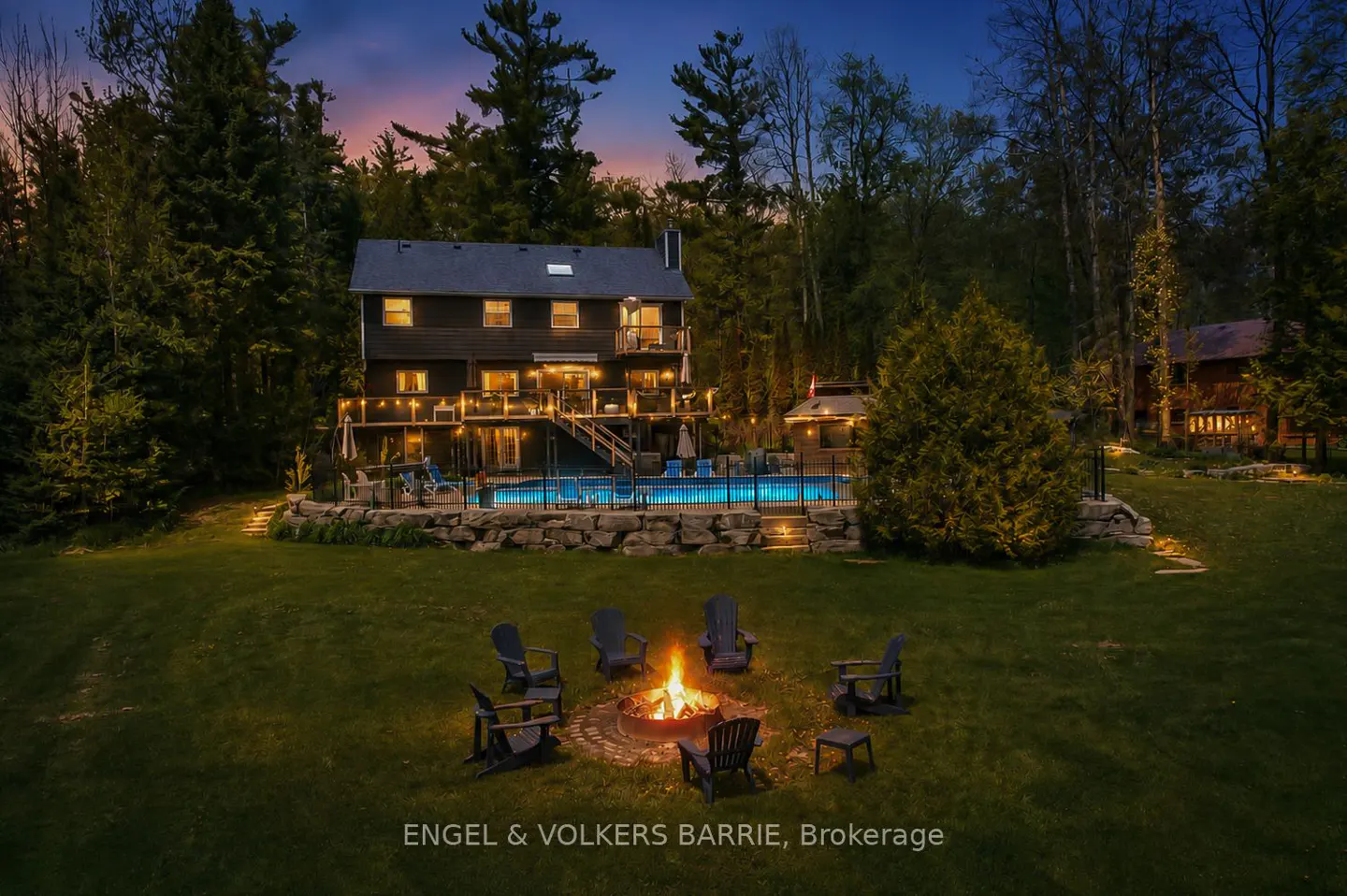 Twilight view of a two-story house with a pool, fire pit, and surrounding trees. The fire pit has chairs around it.