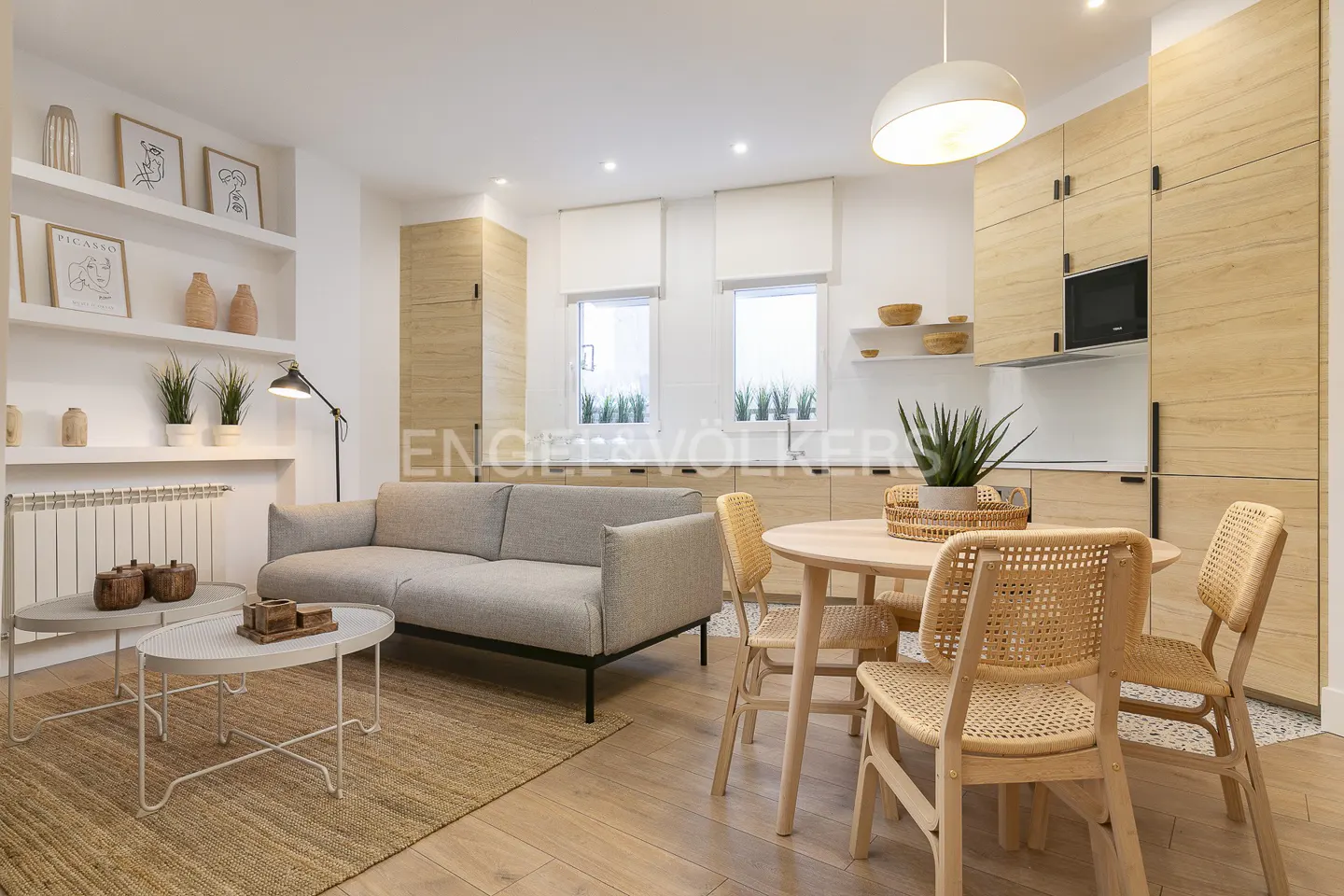 Bright living room with a gray sofa, round table with wicker chairs, and light wood cabinets. Shelves display art and plants.