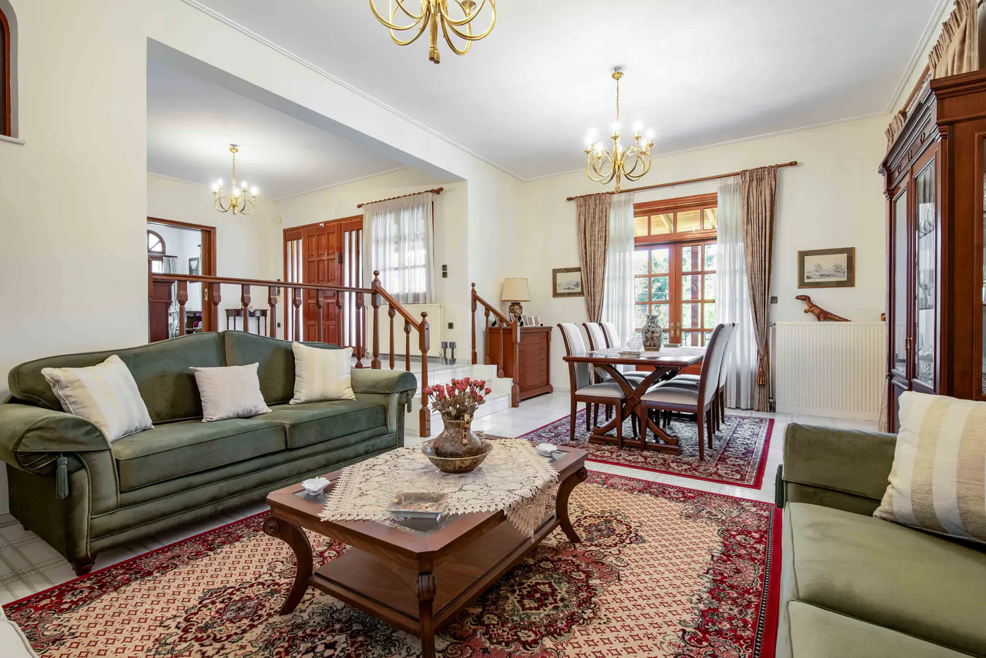 A living room with green sofas, a wooden coffee table, and a dining table with chairs on a patterned rug. Chandeliers hang from the ceiling.