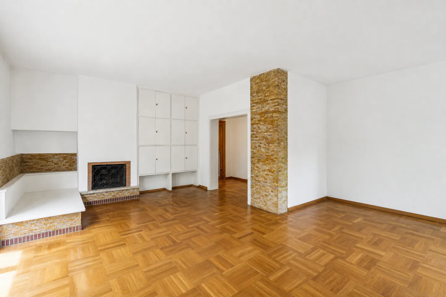 Bright, empty living room with parquet floors, white walls, and a brick fireplace. Built-in white cabinets and a brick pillar add architectural detail.