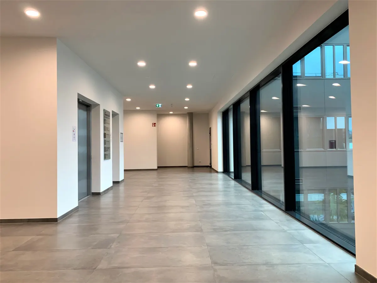 Bright, empty office hallway with gray tile floor and white walls. Large windows line the right side, reflecting the blue sky.