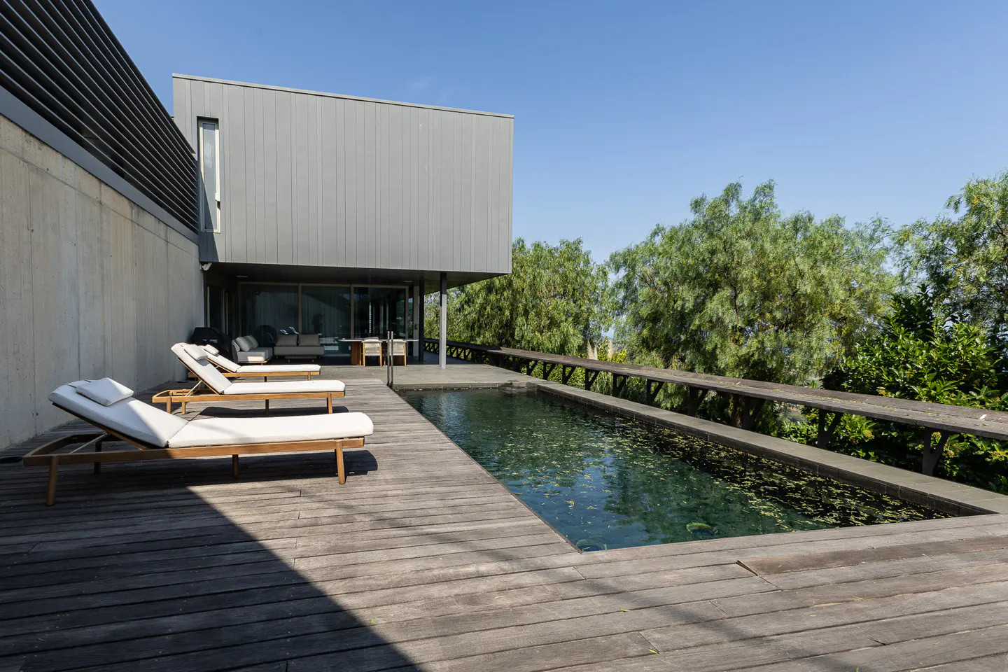 Modern home exterior with a wooden deck, swimming pool, and lounge chairs under a clear blue sky.