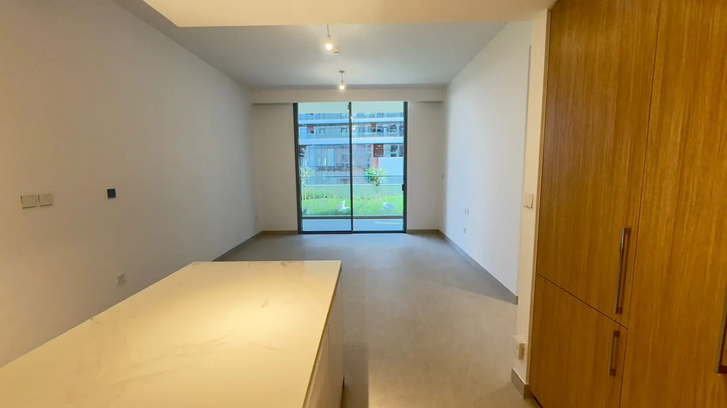 An interior view of a bright, empty apartment with white walls, a sliding glass door to a balcony, and light wood cabinets.
