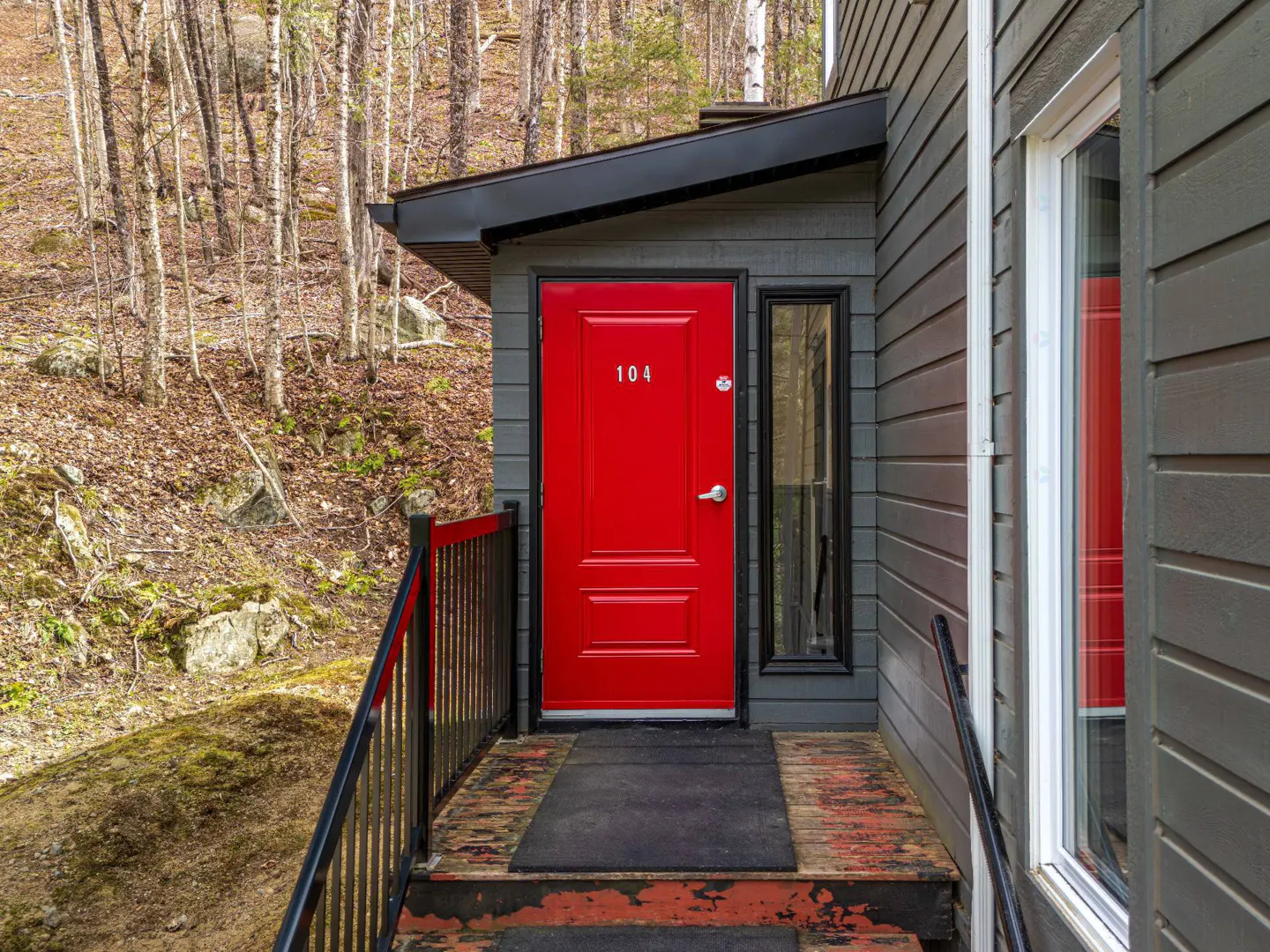 Exterior view of a gray cabin with a bright red door numbered 104, black trim, and a wooded hillside in the background.
