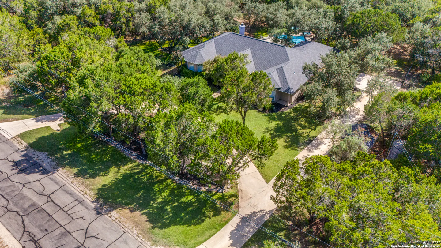 Aerial view of a house with a gray roof, surrounded by lush green trees and a pool in the background.
