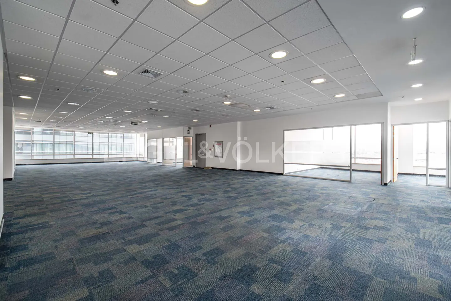 Bright, empty office space with blue patterned carpet and large windows. White walls and ceiling with recessed lighting.
